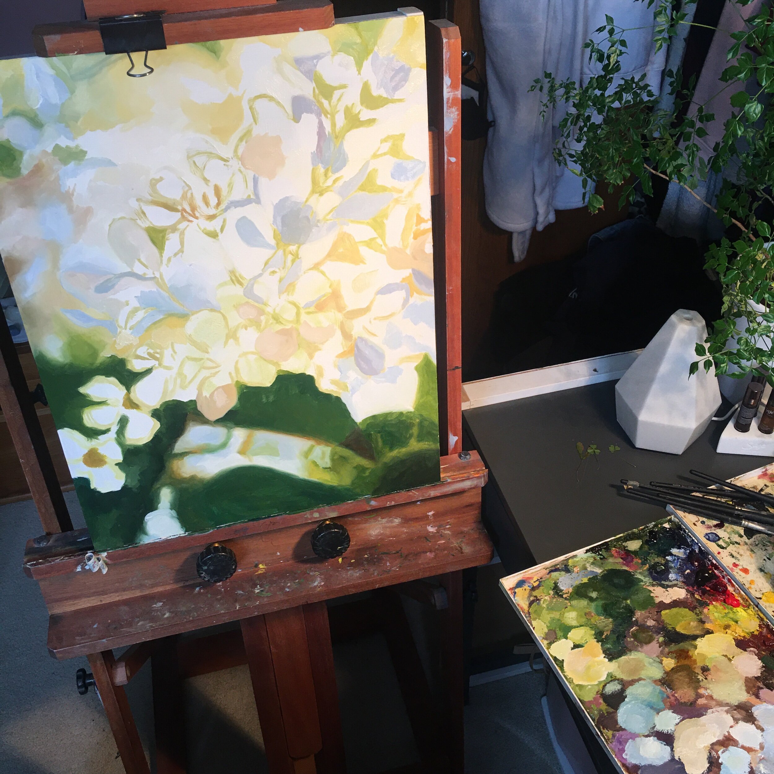A progress photo from a recent painting, enjoying the process. Image description: An unfinished painting of flowers sits on an easel to the left and a colourful palette of paint sits on the corner of a grey desk on the right.