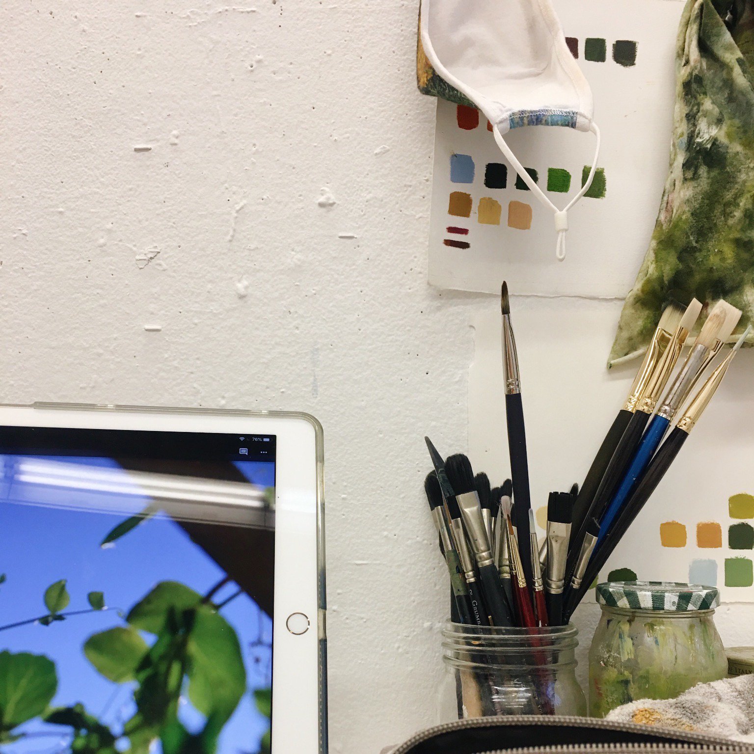 a photo of objects on a desk against a white wall, a corner of an ipad with the picture of a pea plant on it, a jar full of paint brushes next to it, a mask and a piece of paper with color swatches hang on the wall.